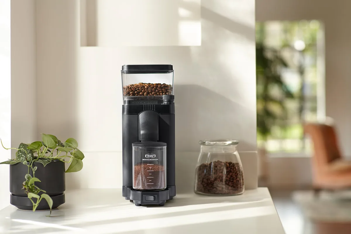 Moccamaster KM5 i matt svart på en lys kjøkkenbenk, ved siden av en potteplante og et glass med bønner