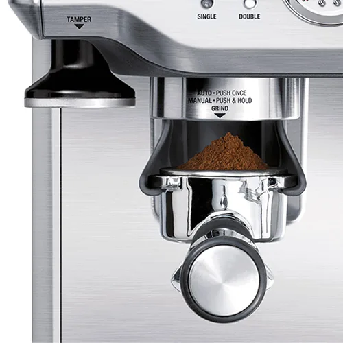Kvernmunningen på Sage Barista Express BES875 med filterholderen full av nymalt kaffe og stamperen til venstre